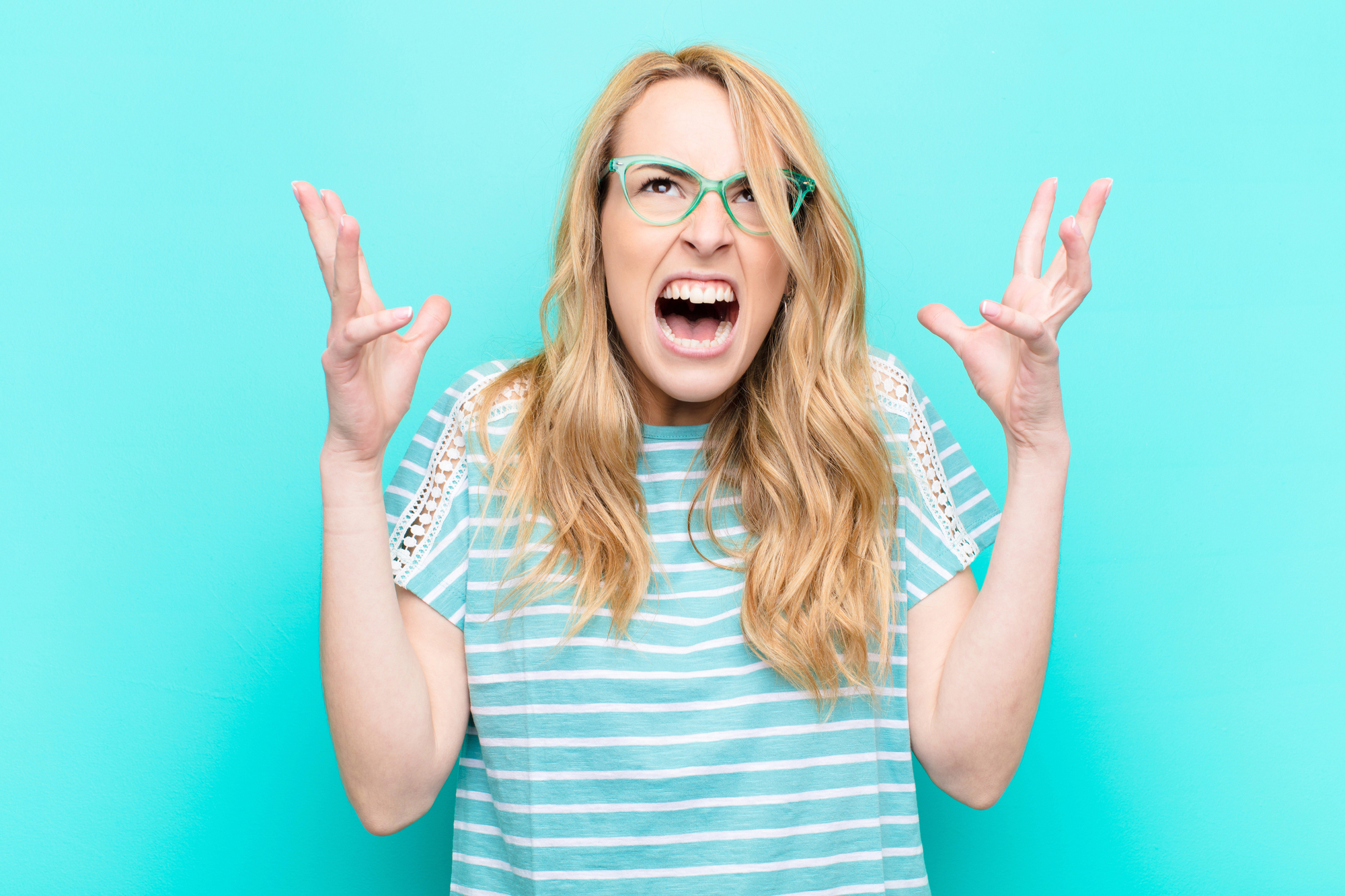 A woman with long blonde hair and green glasses is standing against a turquoise background. She's wearing a striped shirt and has a frustrated expression with her hands raised near her head.