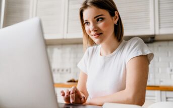 A woman with short hair wearing a white t-shirt is sitting at a table, using a laptop. She is smiling and looking at the screen. The background shows a kitchen with white cabinetry.