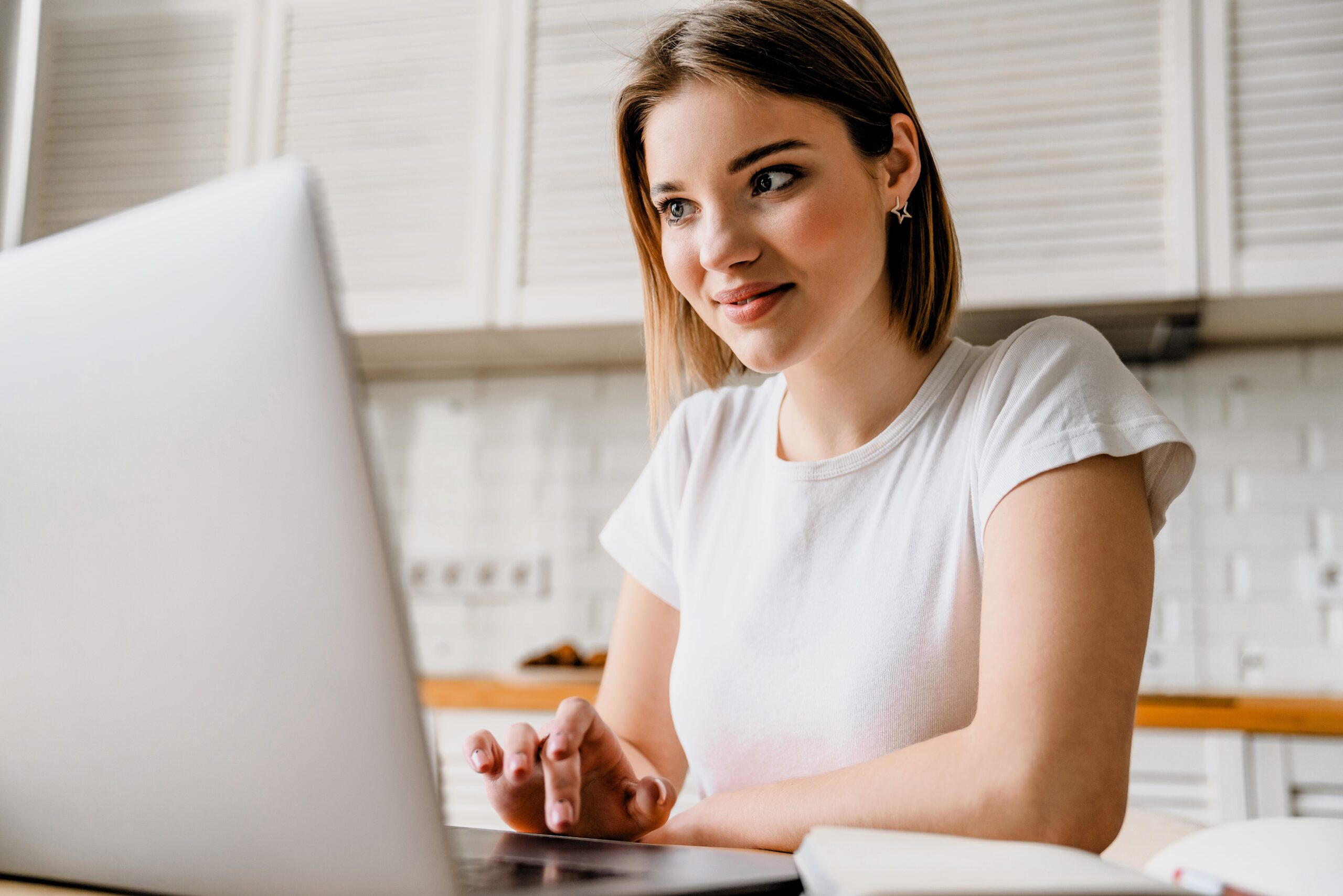 A woman with short hair wearing a white t-shirt is sitting at a table, using a laptop. She is smiling and looking at the screen. The background shows a kitchen with white cabinetry.