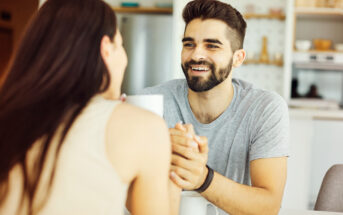 A bearded man smiling while holding hands with a woman at a table. Both have mugs in front of them. The background shows a modern kitchen setting. The atmosphere appears warm and friendly.