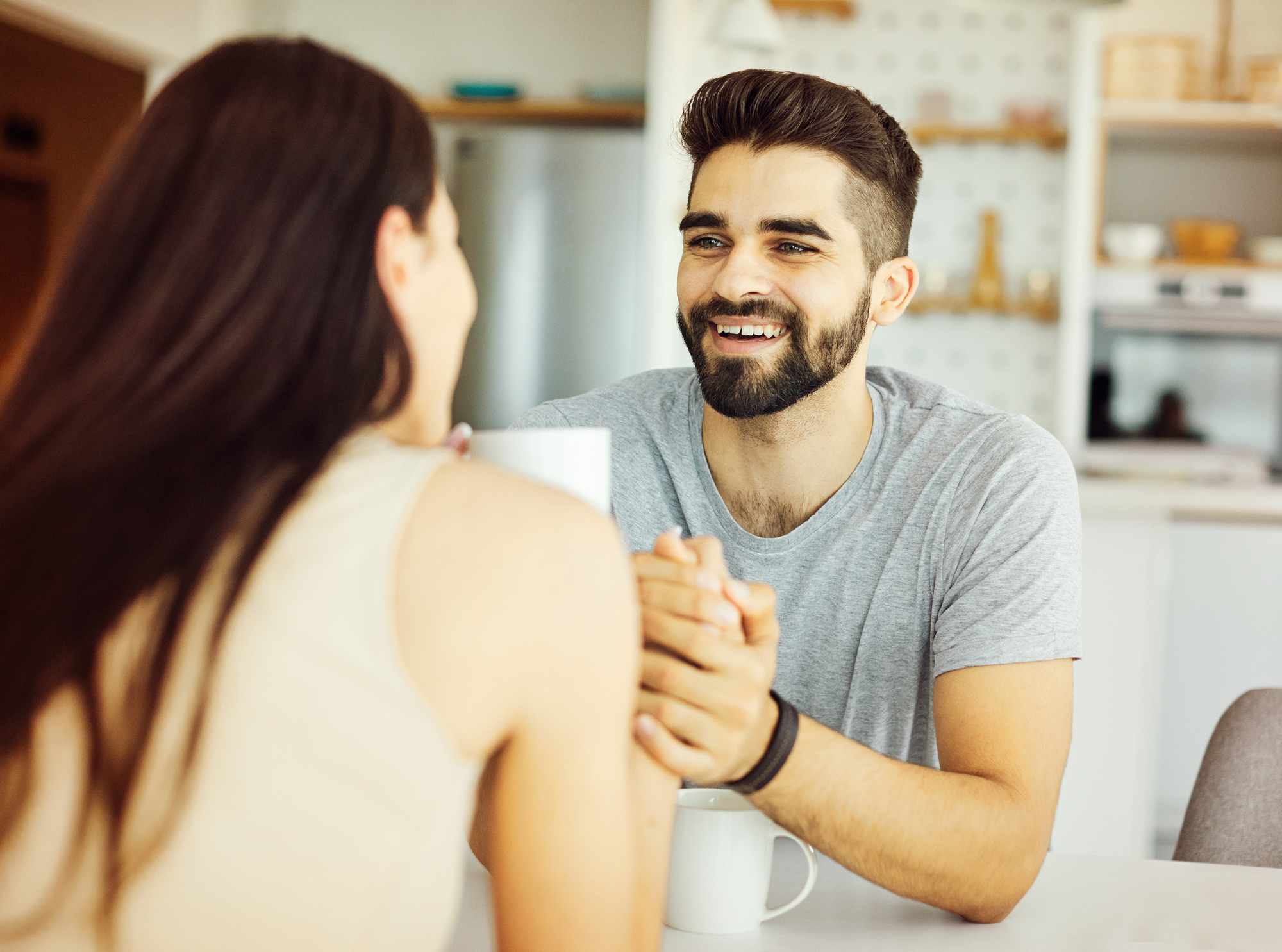 A bearded man smiling while holding hands with a woman at a table. Both have mugs in front of them. The background shows a modern kitchen setting. The atmosphere appears warm and friendly.