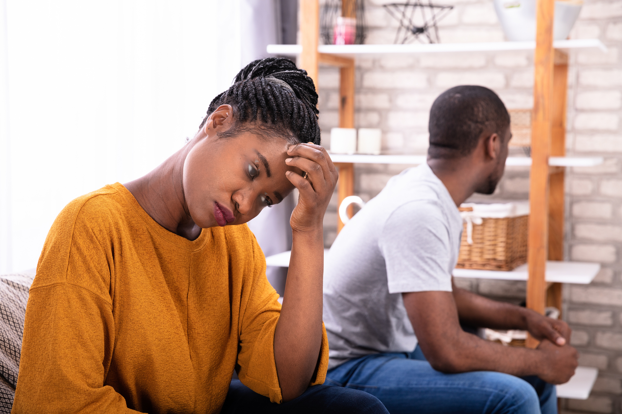 A woman in a yellow sweater sits with her hand on her forehead, looking upset. A man in a gray t-shirt sits beside her, turned away. They are in a cozy room with shelves and a basket in the background.