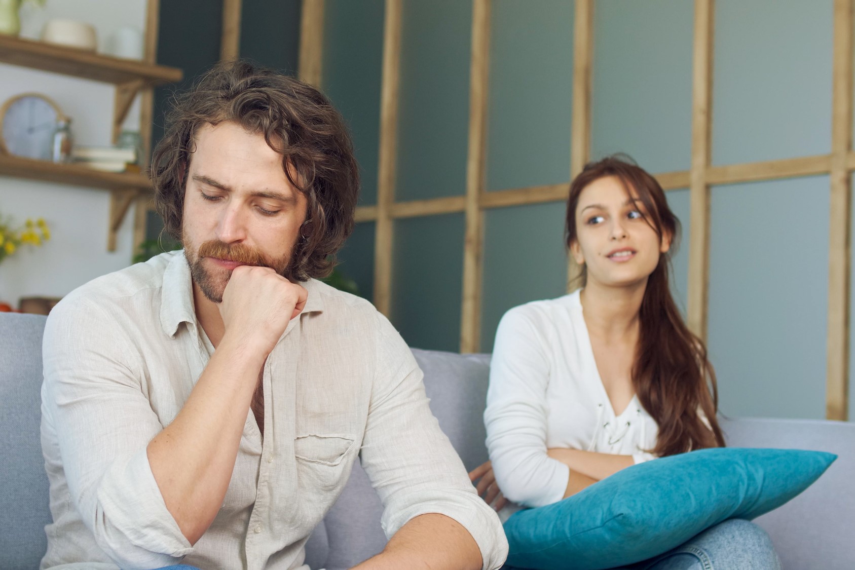 A man with a thoughtful expression sits on a couch, resting his chin on his hand, wearing a light shirt. A woman in a white top sits behind him, leaning back with her arms crossed, holding a blue cushion. The setting is a cozy living room.