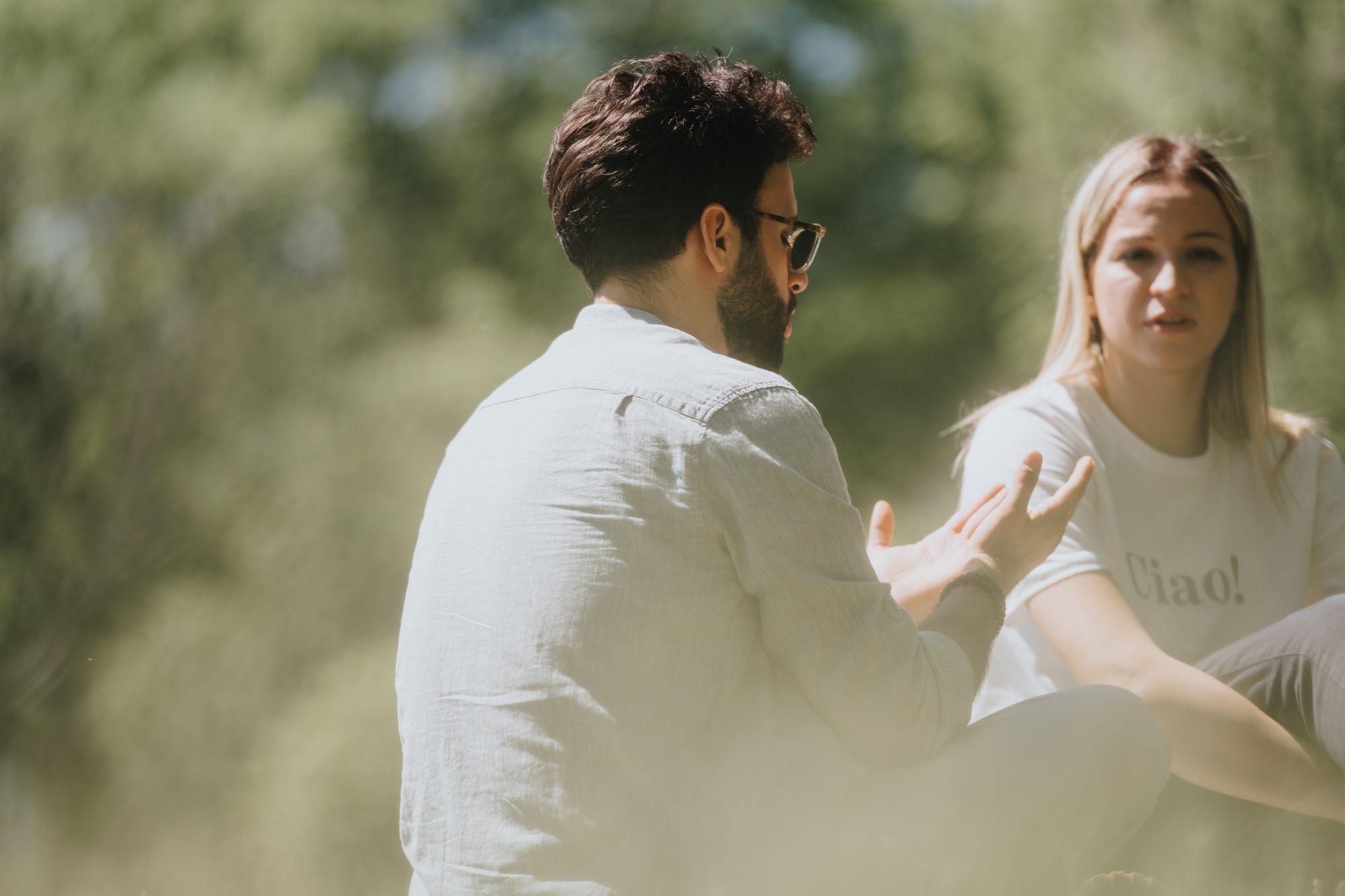 A man and a woman are sitting outdoors on a sunny day, engaged in conversation. The man, wearing a light shirt and sunglasses, gestures with his hands. The woman listens attentively, wearing a white T-shirt with "Ciao!" printed on it. Greenery surrounds them.