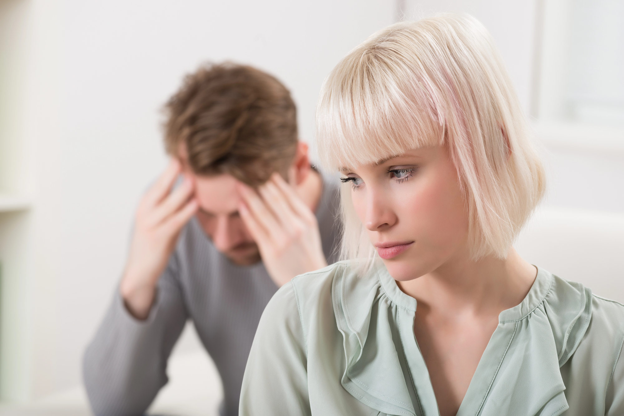 A woman with short blonde hair looks away with a pensive expression. In the background, a man with brown hair sits with his hands on his head, appearing stressed. They are indoors, and the setting is softly lit.