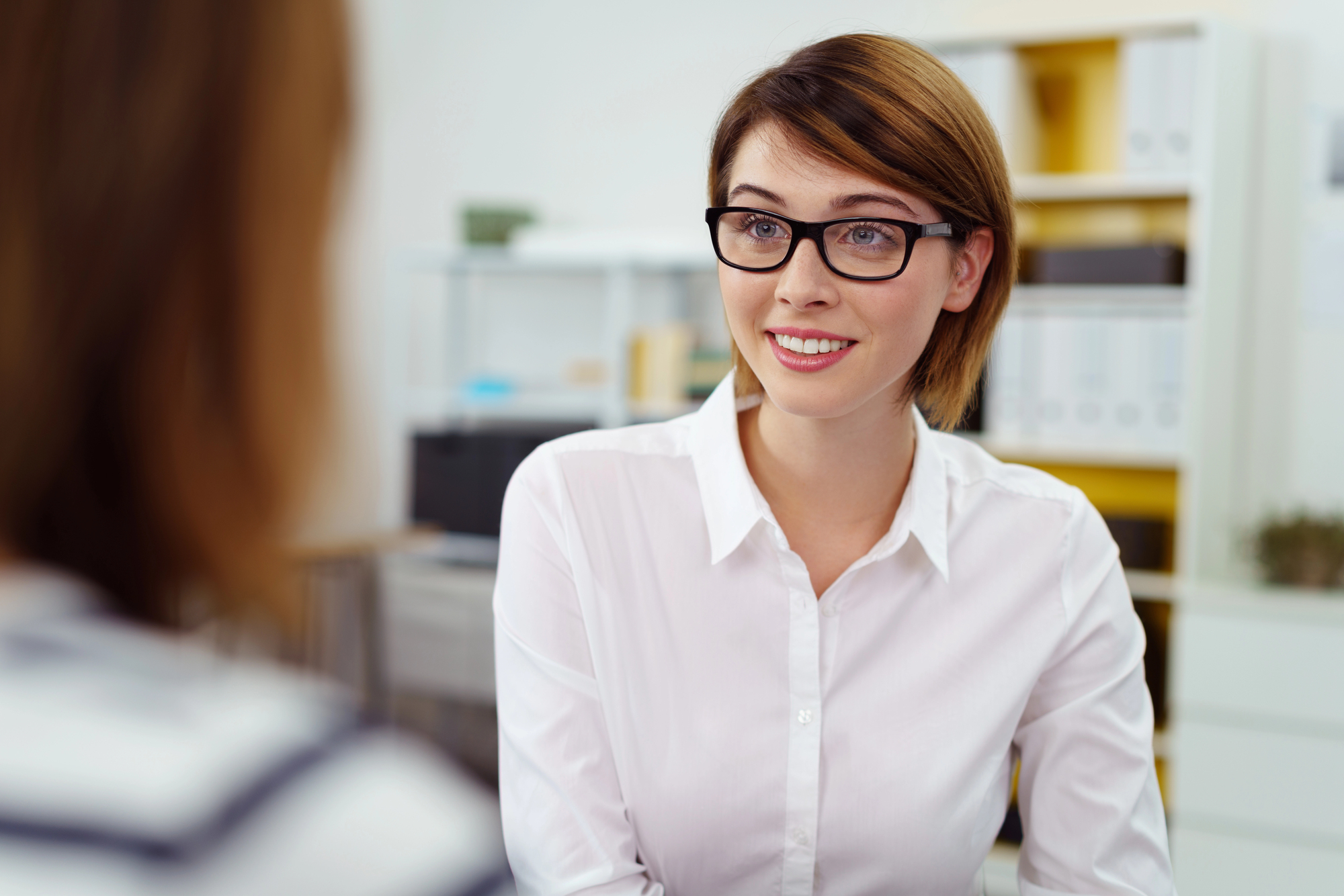 A woman with short brown hair and glasses is smiling while sitting at a desk. She is wearing a white shirt and appears to be talking to another person whose back is visible. The setting is an office with shelves and files in the background.