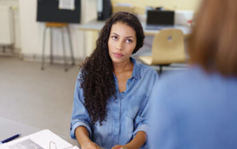 A woman with long curly hair wearing a blue denim shirt sits at a table with papers in front of her. She looks thoughtfully at someone off-camera. The room is blurred in the background, with a chair and a board visible.