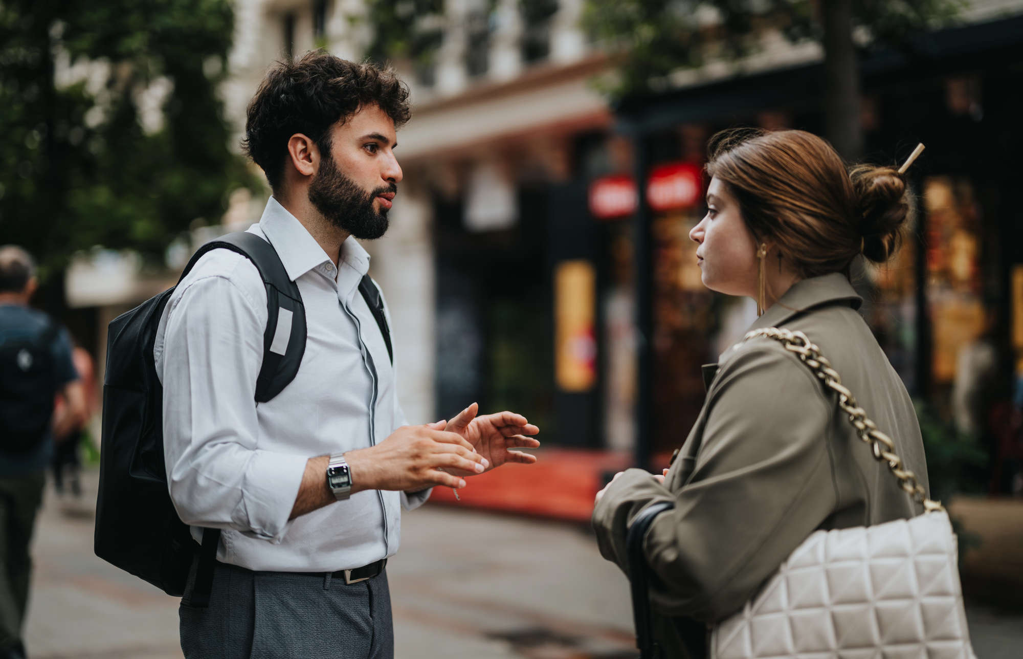 A man with a beard and a backpack engages in conversation with a woman wearing a coat and carrying a white quilted bag. They are standing on a city street with shops and greenery in the background.