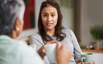 Two people sit on a couch drinking from mugs. One, a young woman, appears to be speaking or explaining something to the other, an older person whose back is to the camera. The setting is a cozy, well-lit living room.