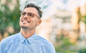 A smiling man with short curly hair and glasses, wearing a light blue button-up shirt, looks upwards. The background is softly blurred with greenery and buildings, suggesting an outdoor setting on a sunny day.