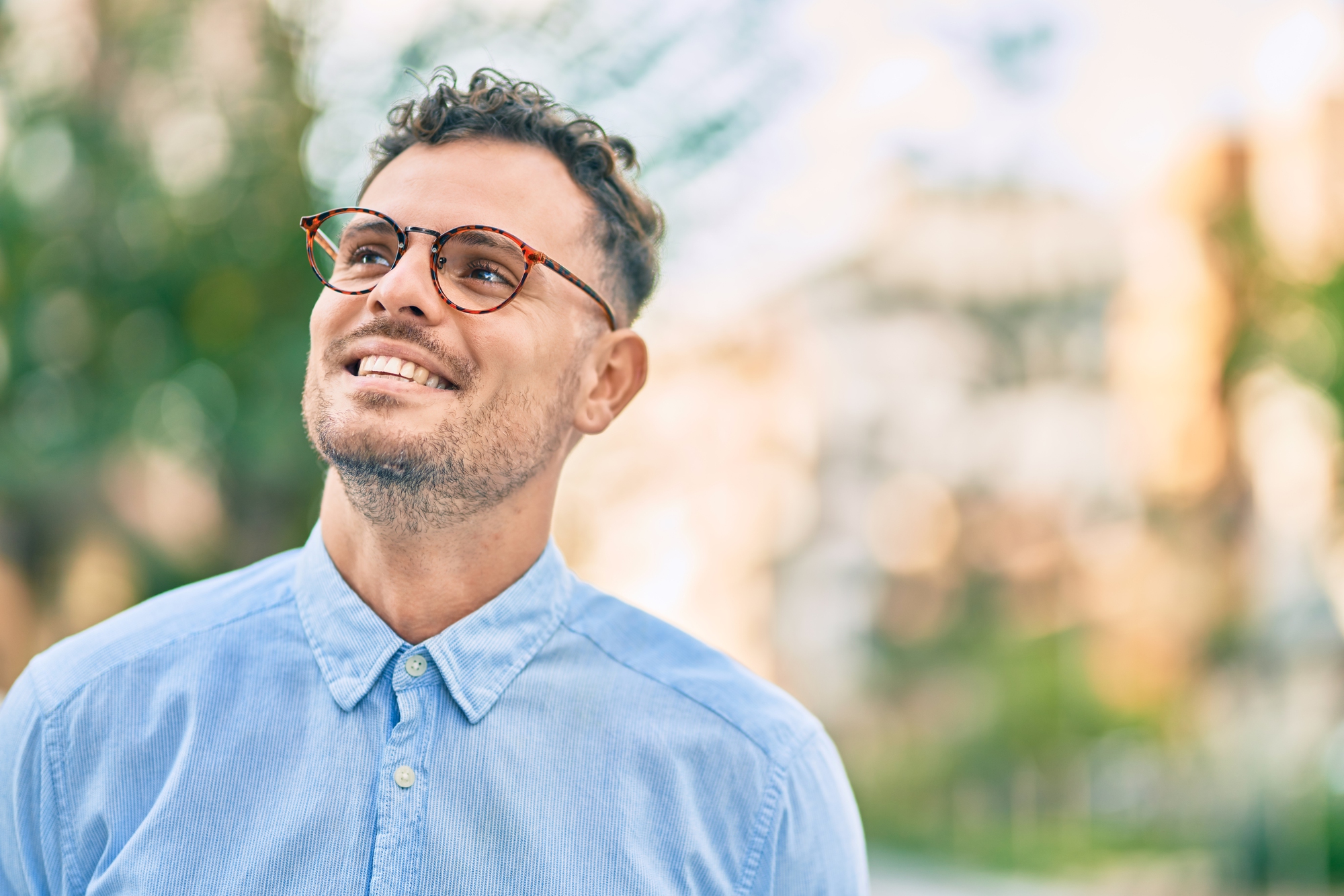 A smiling man with short curly hair and glasses, wearing a light blue button-up shirt, looks upwards. The background is softly blurred with greenery and buildings, suggesting an outdoor setting on a sunny day.