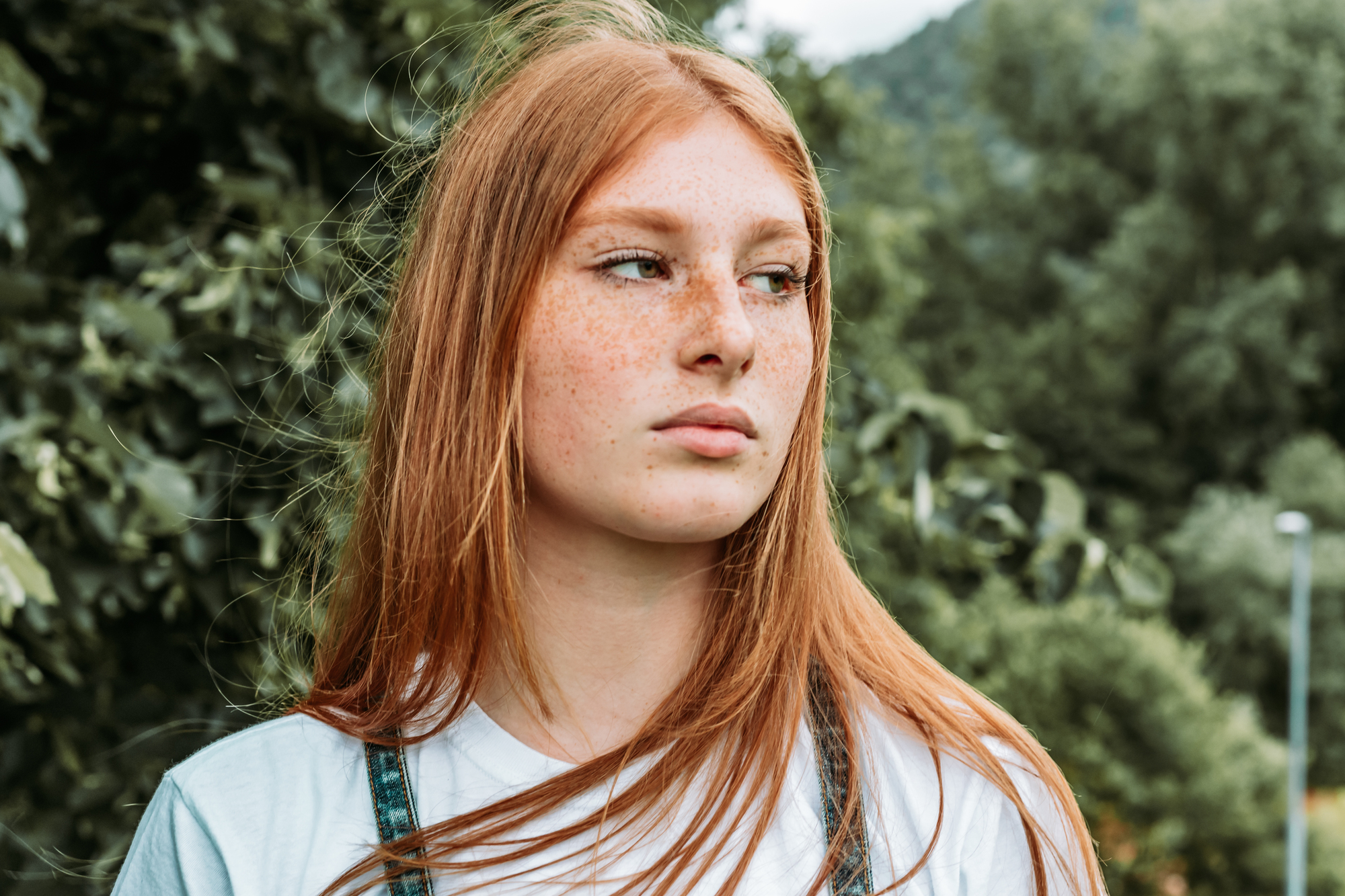 A young person with long red hair and freckles stands outdoors, wearing a white shirt and denim overalls. They are gazing off to the side, with a backdrop of lush green trees and a cloudy sky.