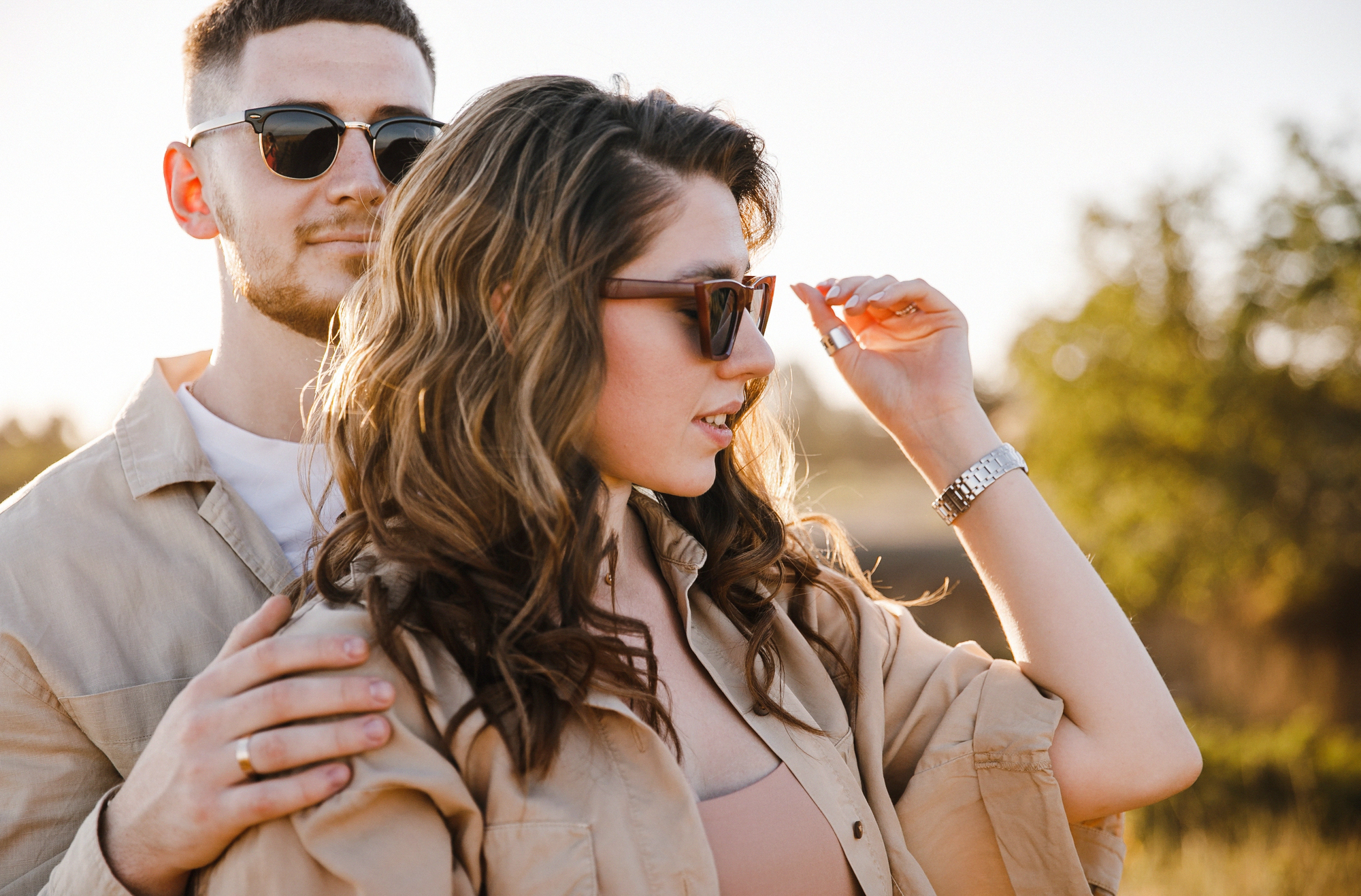 A man and a woman wearing sunglasses stand together outdoors, with the woman adjusting her sunglasses. The sun casts a warm glow, and they are surrounded by a blurred natural background. Both wear light-colored clothing and appear relaxed.