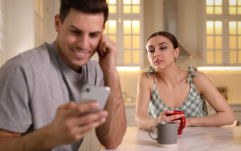 A woman in a green checkered top drinks from a red mug while looking at a man in a gray shirt who is focused on his smartphone. They are sitting at a kitchen table, and the room is softly lit with warm light from the windows.