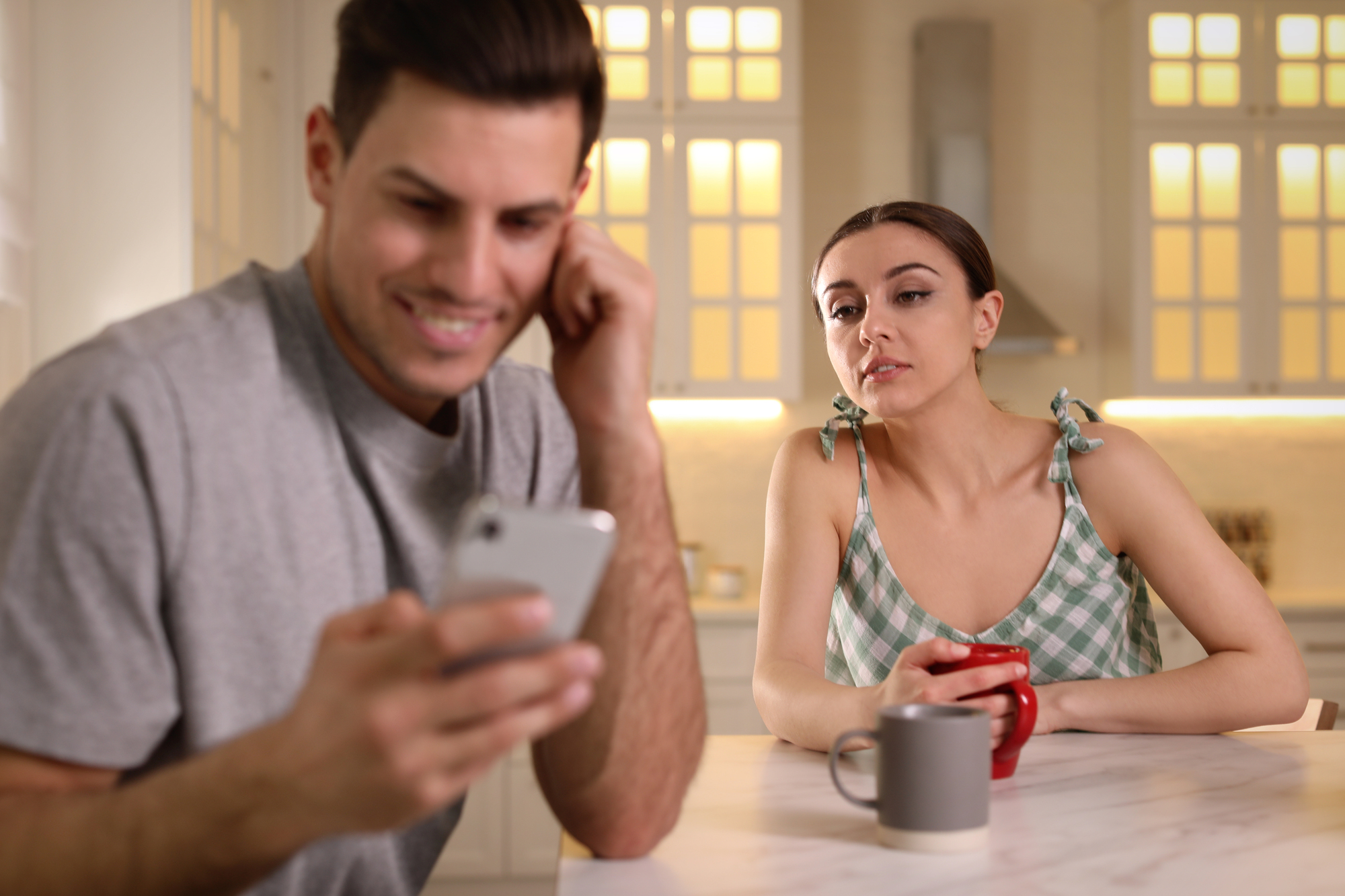 A woman in a green checkered top drinks from a red mug while looking at a man in a gray shirt who is focused on his smartphone. They are sitting at a kitchen table, and the room is softly lit with warm light from the windows.