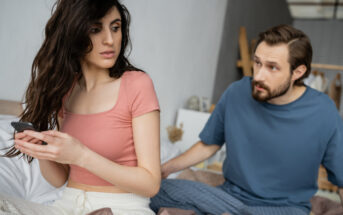 A woman with long dark hair looks concerned while holding a phone, sitting on a bed. A man with short hair and a beard, wearing a blue shirt, sits beside her, appearing worried and leaning towards her. The setting is a softly lit bedroom.