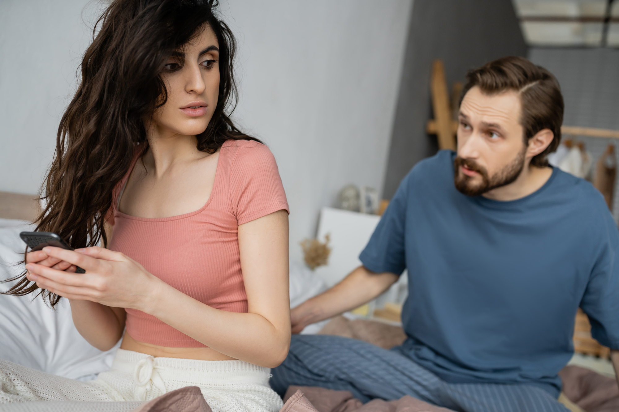 A woman with long dark hair looks concerned while holding a phone, sitting on a bed. A man with short hair and a beard, wearing a blue shirt, sits beside her, appearing worried and leaning towards her. The setting is a softly lit bedroom.