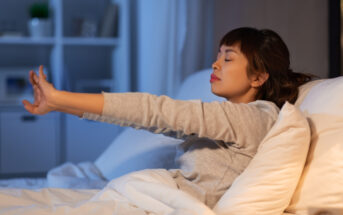 A woman stretches her arms forward while sitting up in bed. She looks relaxed with her eyes closed, wearing a long-sleeve top. The room is softly lit, creating a calm atmosphere.