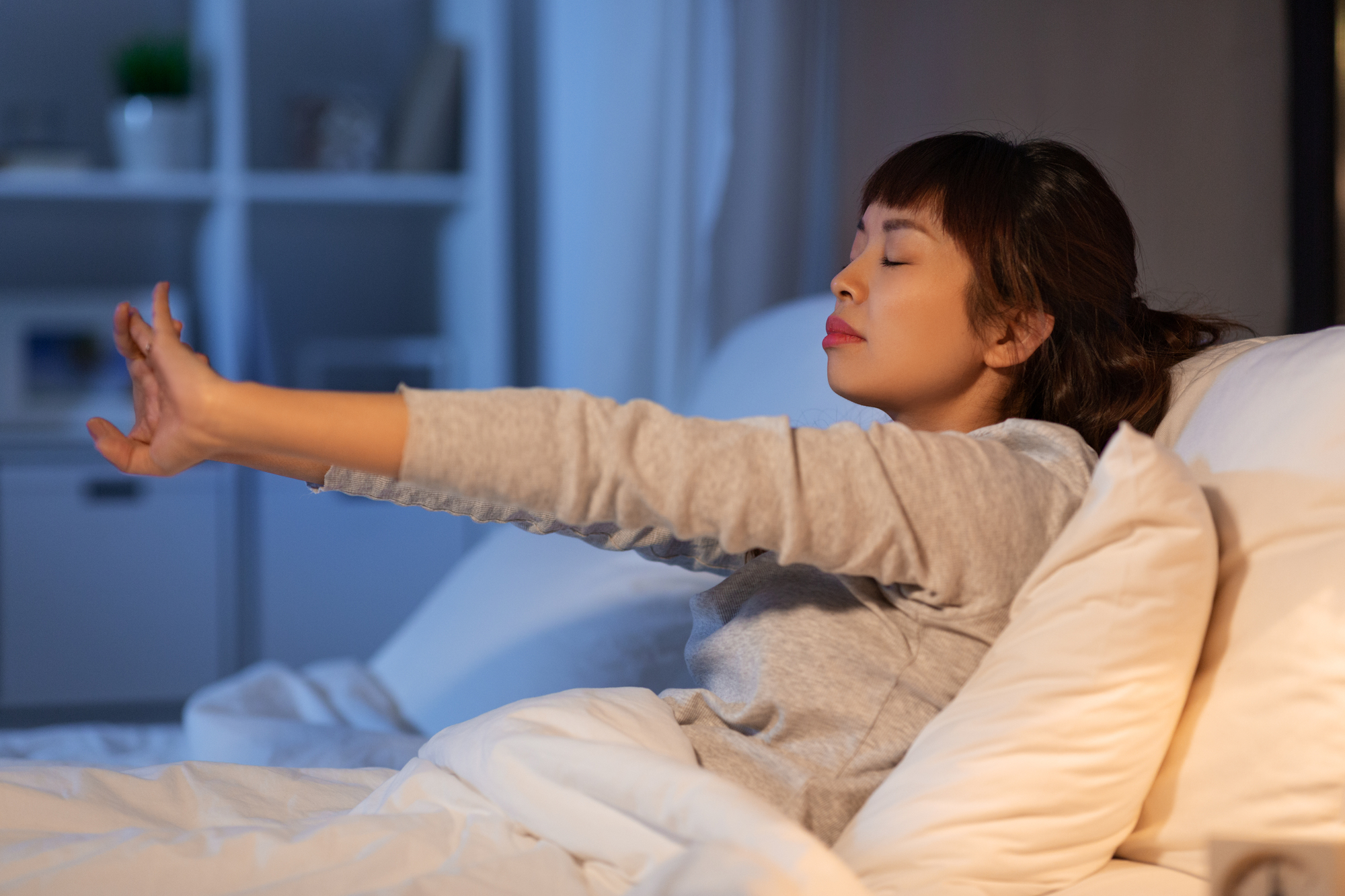 A woman stretches her arms forward while sitting up in bed. She looks relaxed with her eyes closed, wearing a long-sleeve top. The room is softly lit, creating a calm atmosphere.