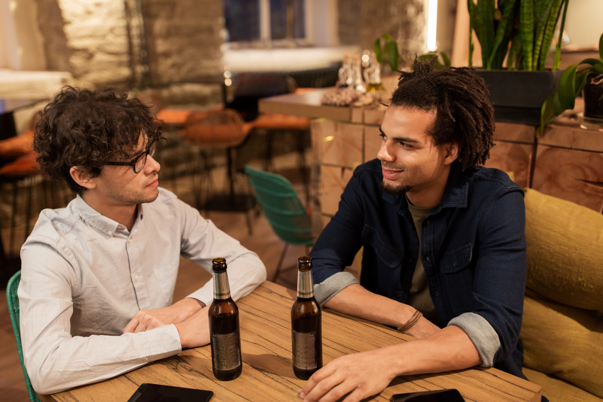 Two men are sitting at a wooden table in a cozy cafe, engaged in conversation. Each has a bottle of beer in front of them. The atmosphere is warm, with soft lighting and comfortable seating in the background.