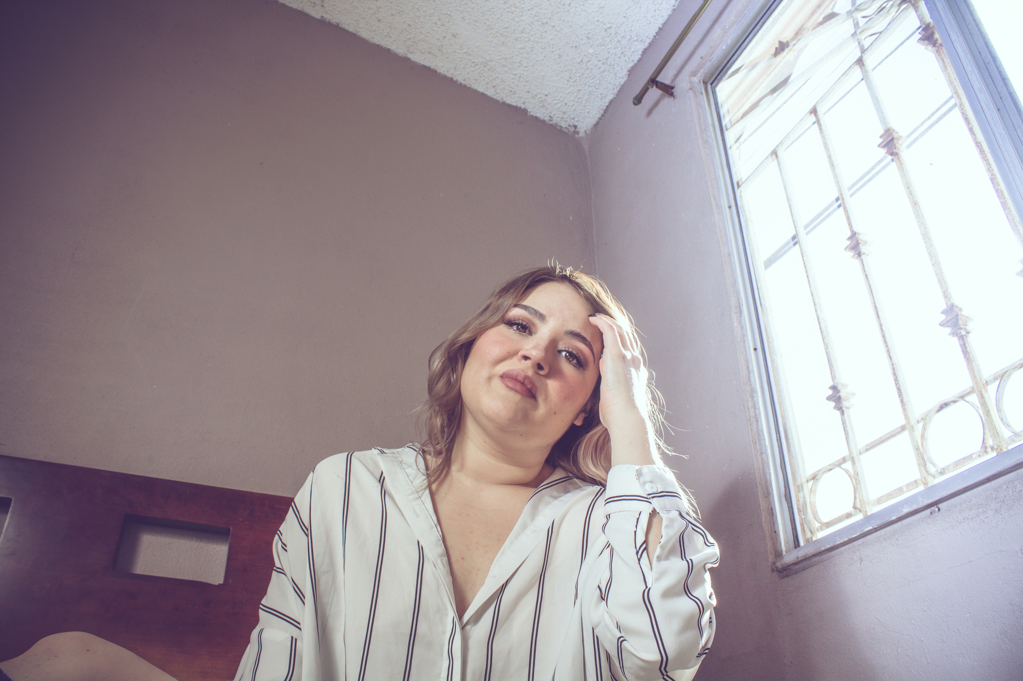 A woman with shoulder-length hair and a striped shirt sits on a bed, resting her head on her hand. She is smiling slightly, with a window and soft light in the background.