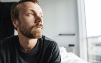 A man with a beard and mustache gazes thoughtfully out of a window. He is wearing a dark shirt and sitting in a room with soft natural light filtering in. The mood appears contemplative.