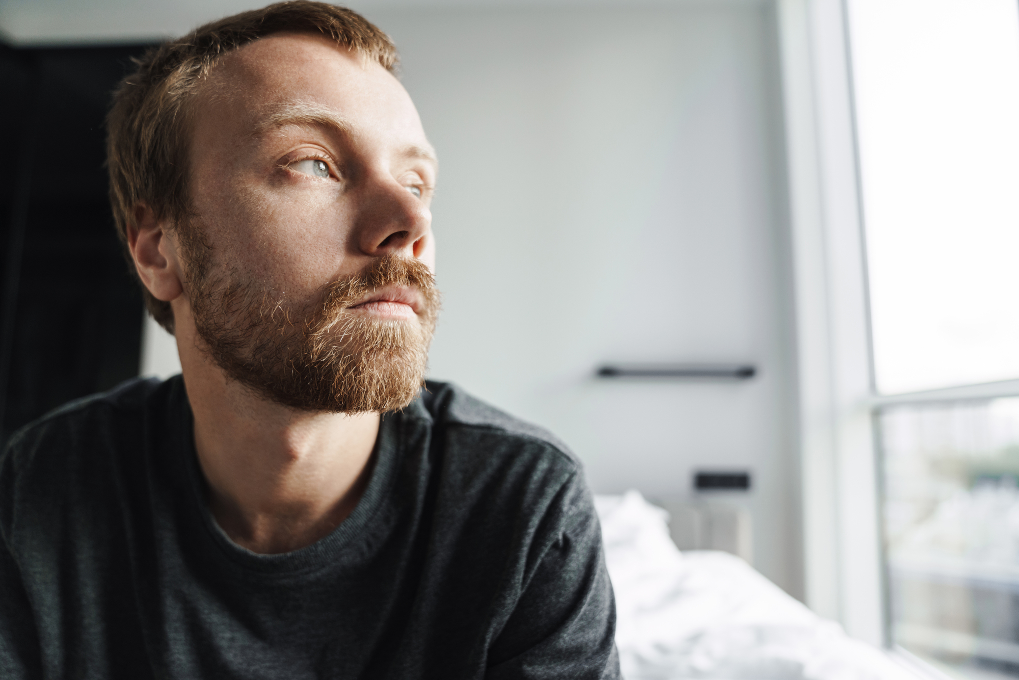 A man with a beard and mustache gazes thoughtfully out of a window. He is wearing a dark shirt and sitting in a room with soft natural light filtering in. The mood appears contemplative.