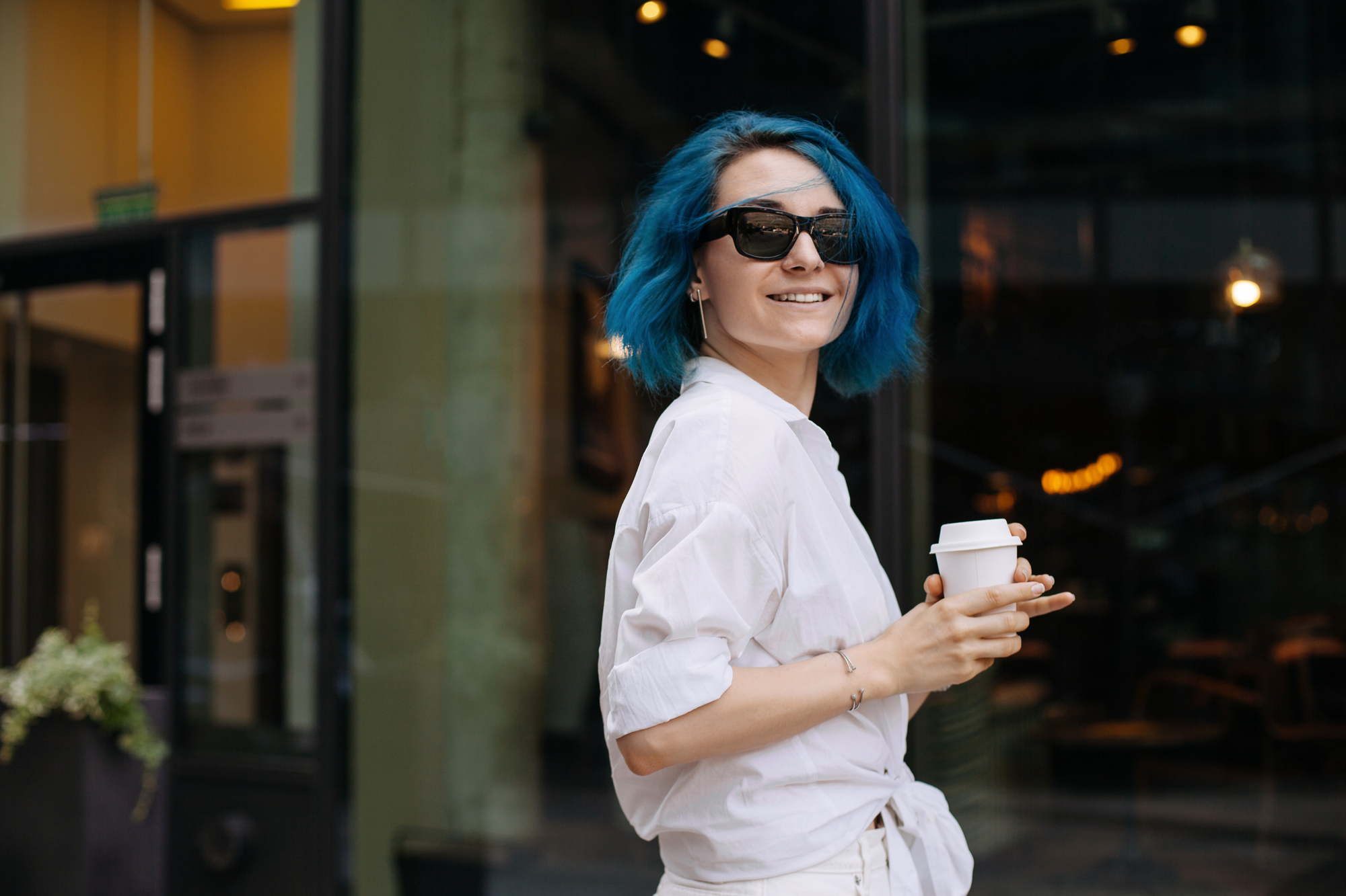 A person with bright blue hair, wearing sunglasses and a white outfit, holds a coffee cup while standing outside a building. The glass door in the background reflects outdoor light, and a potted plant is visible on the left.