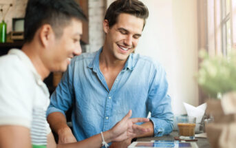 Two people sit at a table, smiling and looking at a tablet. One person wears a blue shirt and the other a striped shirt. There's a glass of iced coffee and a stack of napkins on the table by a window.