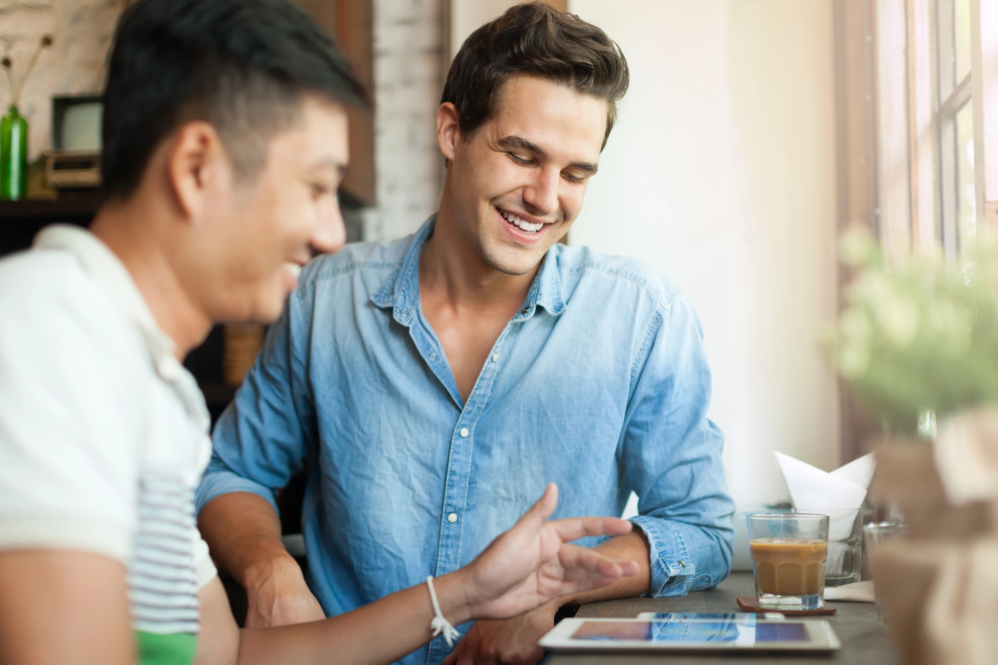 Two people sit at a table, smiling and looking at a tablet. One person wears a blue shirt and the other a striped shirt. There's a glass of iced coffee and a stack of napkins on the table by a window.