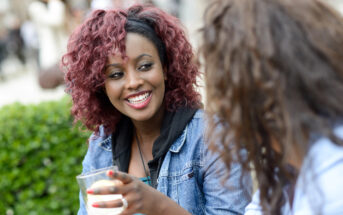 Two people sitting outdoors, engaging in conversation. One is smiling widely while holding a drink. They are surrounded by greenery, creating a relaxed and pleasant atmosphere.