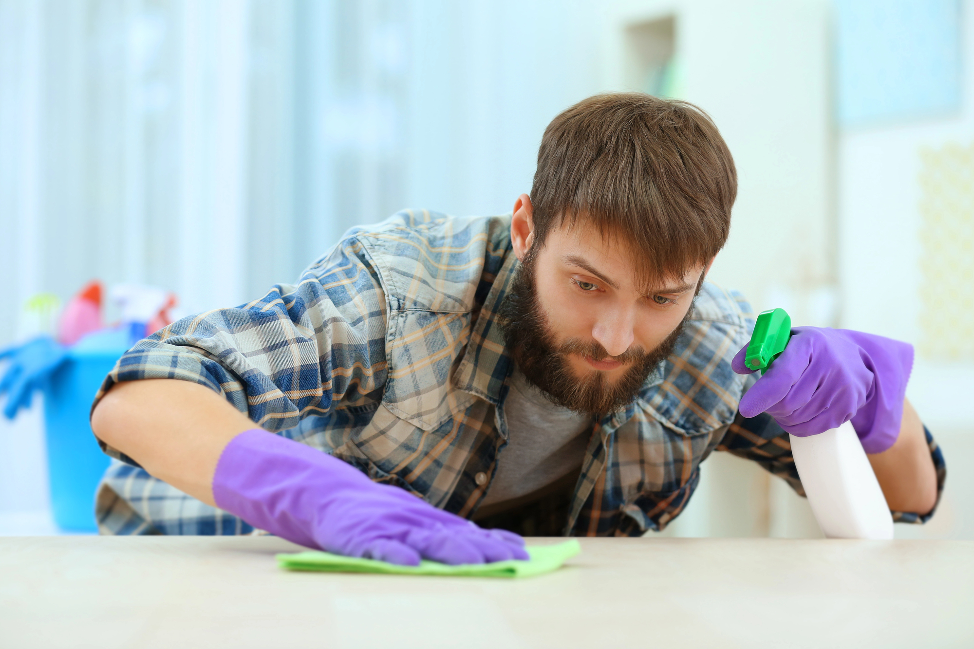 A bearded man wearing a plaid shirt and purple gloves is intently cleaning a surface with a cloth in one hand and a spray bottle in the other. The background is blurred, showing household cleaning supplies.