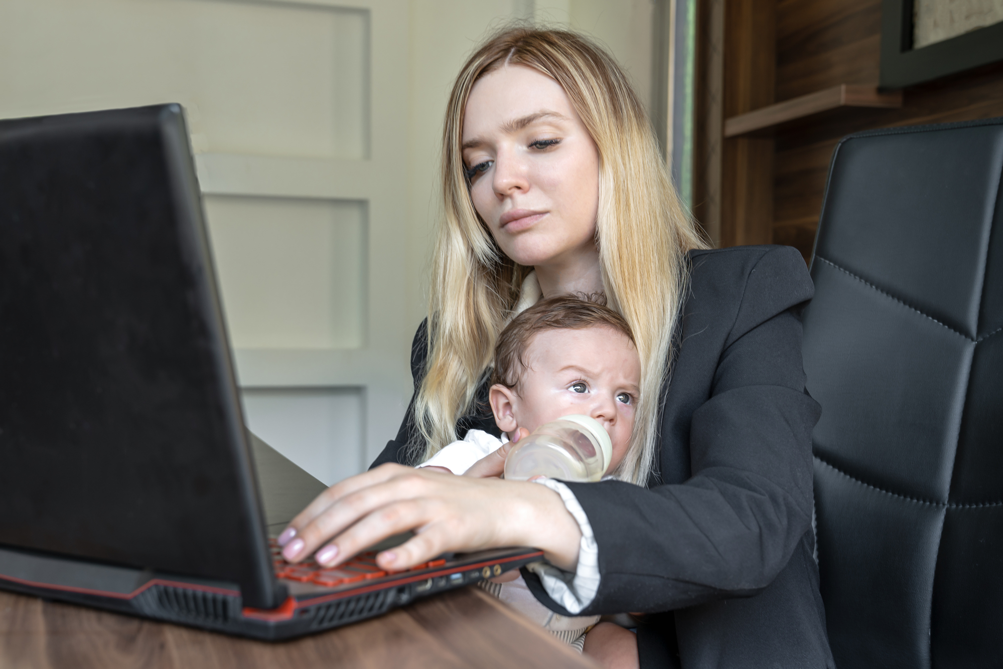 A woman in business attire works on a laptop while holding a baby. The baby is drinking from a bottle. They are seated at a desk within a modern home office setting.