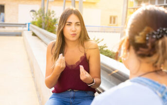 A woman in a red tank top and jeans gestures with her hands while talking to another person on a sunny patio. The background features a metal railing and a building.