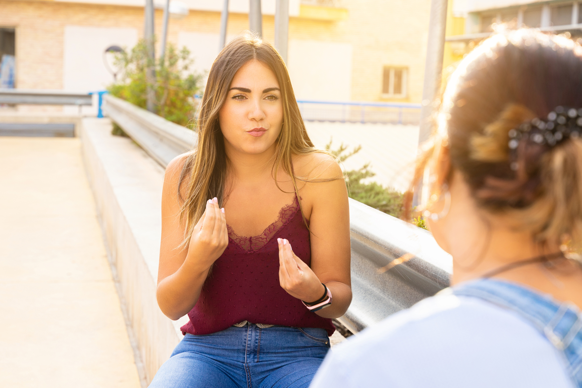 A woman in a red tank top and jeans gestures with her hands while talking to another person on a sunny patio. The background features a metal railing and a building.