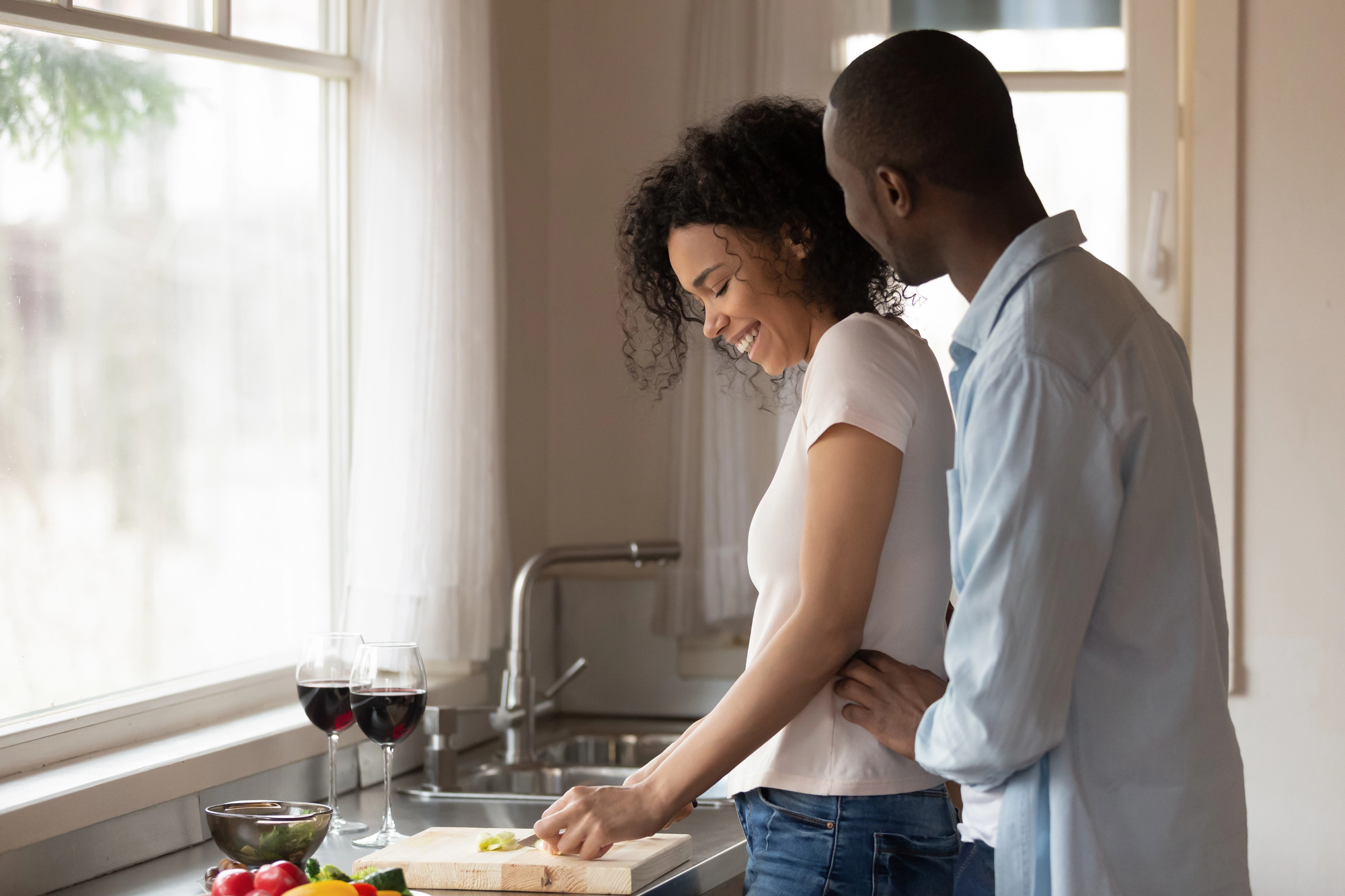 A couple happily cooking together in a kitchen. The woman is chopping vegetables on a cutting board, and the man is standing close behind her. Two glasses of red wine are on the counter near fresh produce. Sunlight filters through a nearby window.