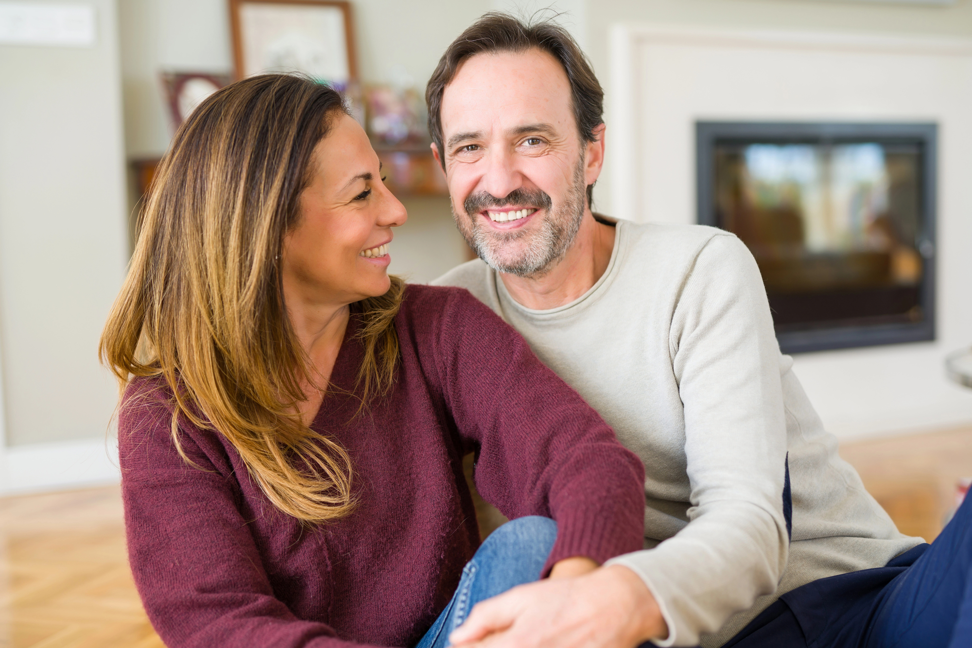 A couple sitting on a floor, smiling at each other. The woman has long hair and is wearing a red sweater, while the man has short hair and is wearing a beige sweater. A fireplace and framed photos are in the background.