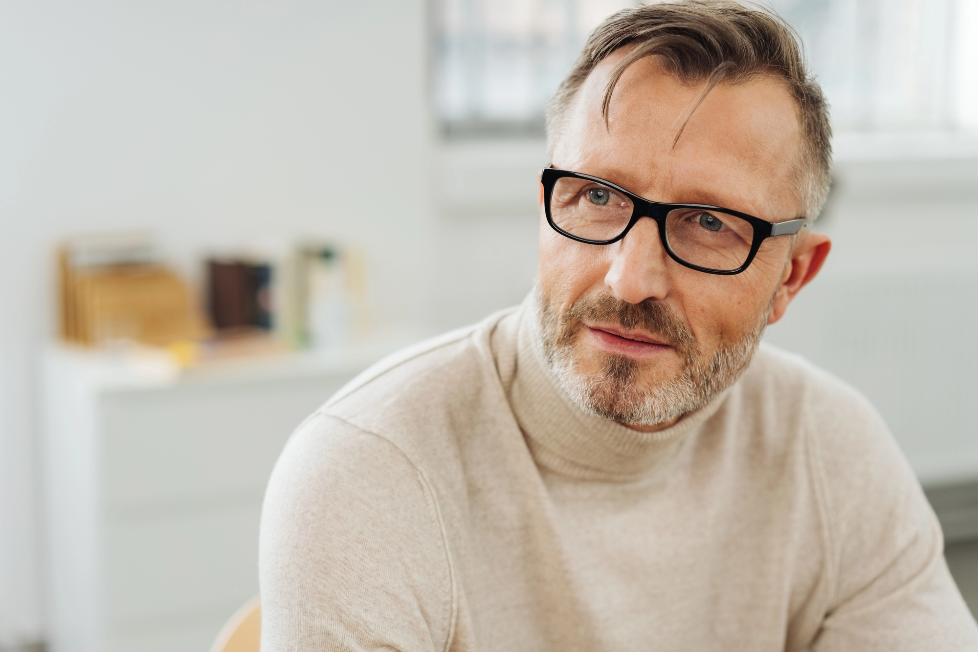 A middle-aged man with glasses and a beard is wearing a light beige turtleneck. He is sitting indoors in a brightly lit room with a soft focus on the background, which includes a white cabinet and some books.