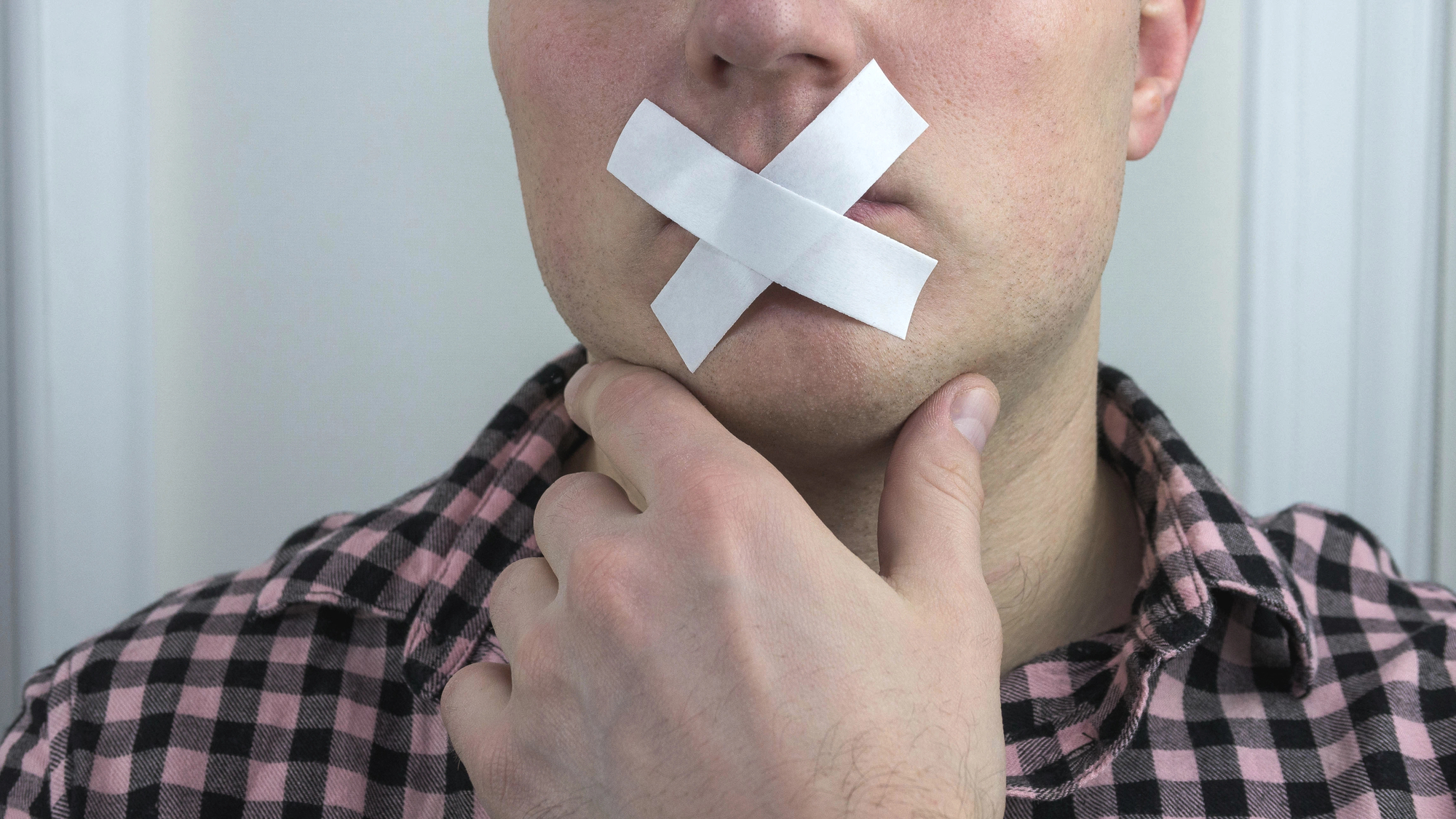 A person wearing a checkered shirt has two pieces of white tape forming an X over their mouth, with one hand gently touching their chin. The image suggests themes of silence or censorship.