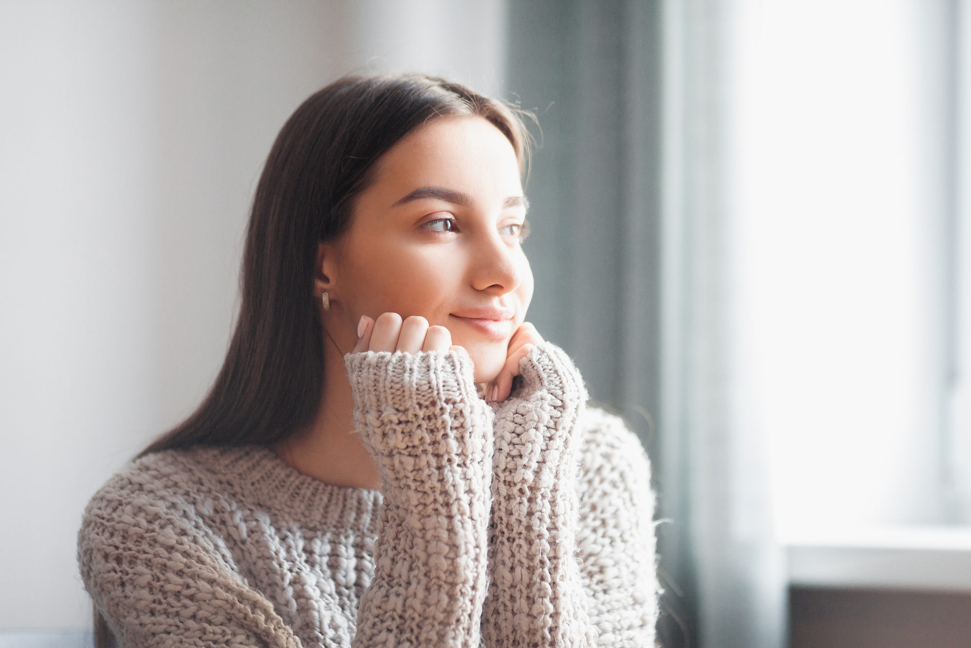 A person with long dark hair wearing a cozy knit sweater sits indoors, resting their chin on their hands, gazing thoughtfully out of a sunlit window. The atmosphere is calm and contemplative.