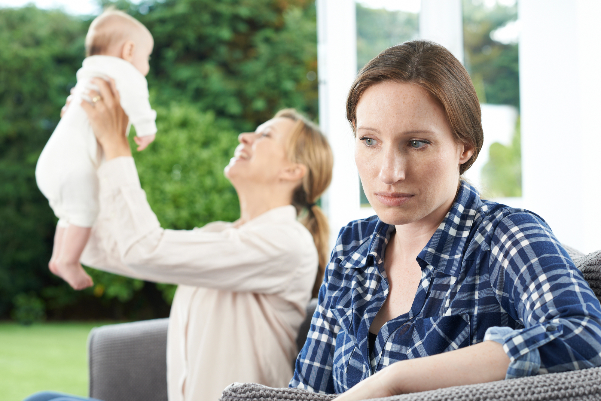 A woman in a plaid shirt sits looking thoughtful on a couch. In the background, another woman joyfully lifts a baby in the air, showing a contrast in emotions. The setting appears to be a bright, cozy living room with a view of greenery outside.