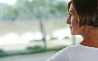 A woman with short brown hair is looking out at a blurred, serene outdoor landscape. She is wearing a white top and appears to be contemplative or relaxed. The background features trees and a body of water.