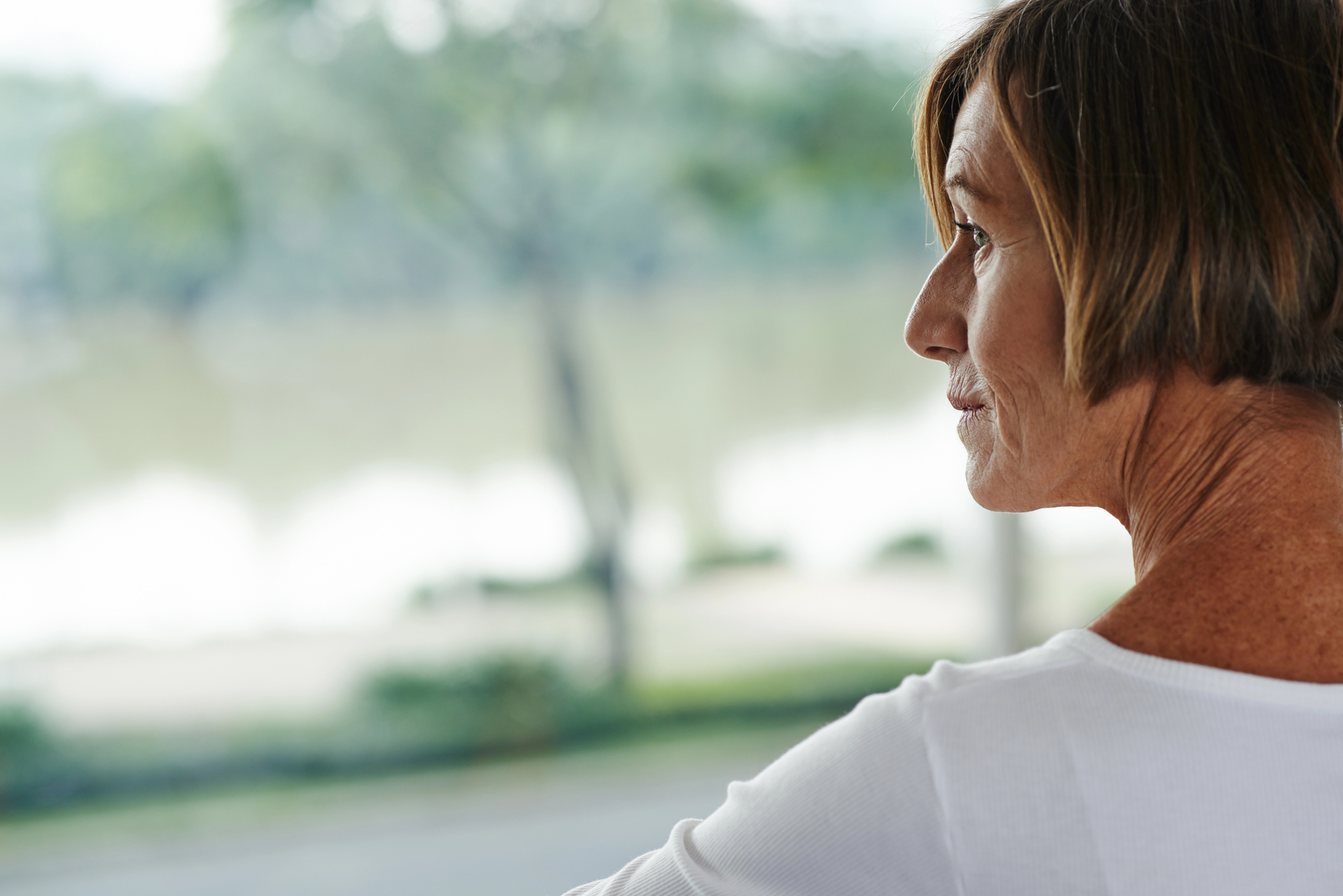 A woman with short brown hair is looking out at a blurred, serene outdoor landscape. She is wearing a white top and appears to be contemplative or relaxed. The background features trees and a body of water.