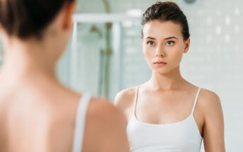 A woman with dark hair tied back is wearing a white tank top and looking at herself in the bathroom mirror. The background has a blurred shower area, creating a soft focus on her reflection.