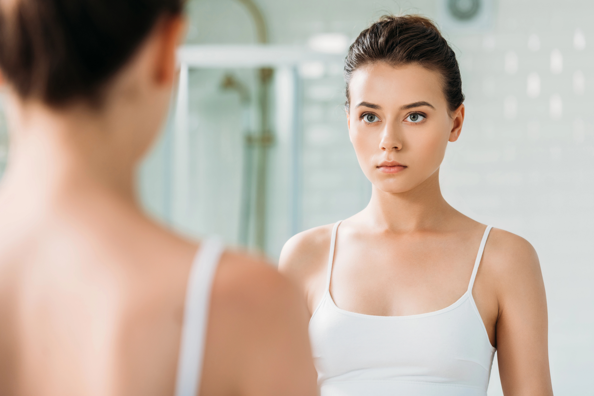 A woman with dark hair tied back is wearing a white tank top and looking at herself in the bathroom mirror. The background has a blurred shower area, creating a soft focus on her reflection.