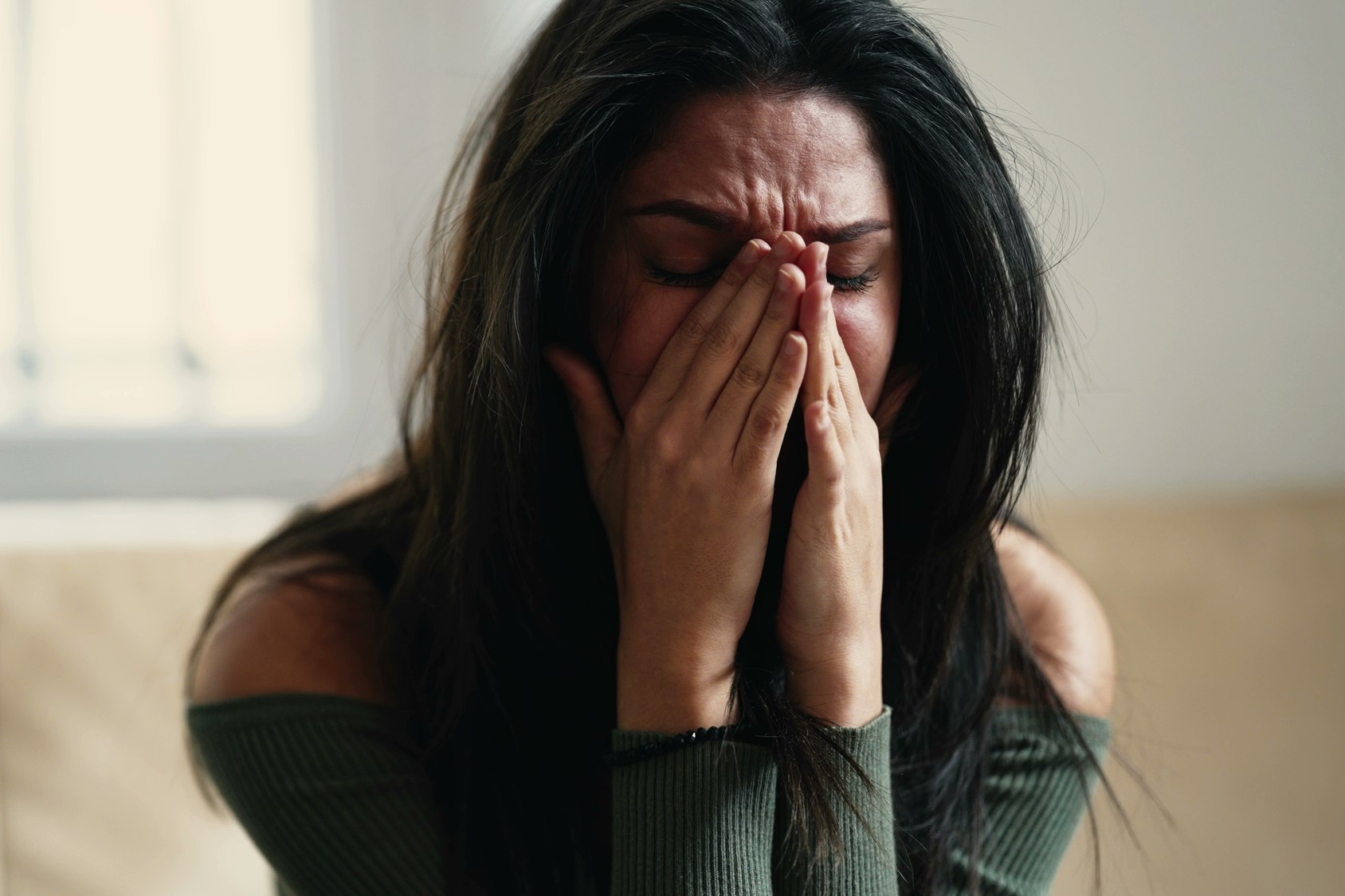 A woman with long dark hair sits inside a room, covering her face with her hands in an expression of distress or sadness. She is wearing a green off-shoulder top and appears to be in a cozy, softly lit environment.