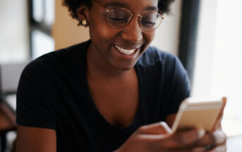 A person wearing glasses and a black T-shirt smiles while looking at a smartphone. They are indoors with soft lighting.