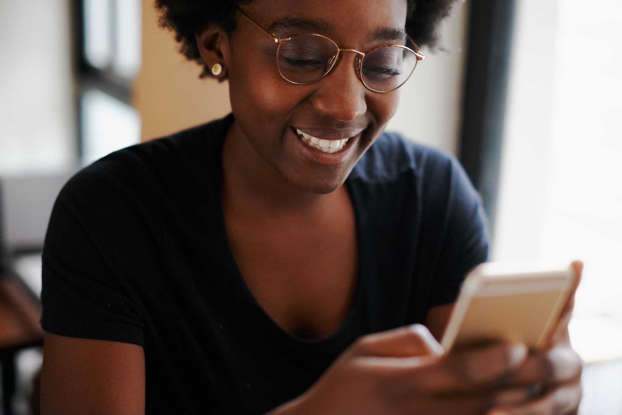 A person wearing glasses and a black T-shirt smiles while looking at a smartphone. They are indoors with soft lighting.