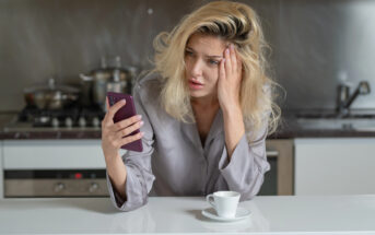 A woman with disheveled blonde hair looks worriedly at her smartphone while sitting at a kitchen counter. She wears a gray shirt and has a white coffee cup in front of her. In the background, there are kitchen appliances and pans.