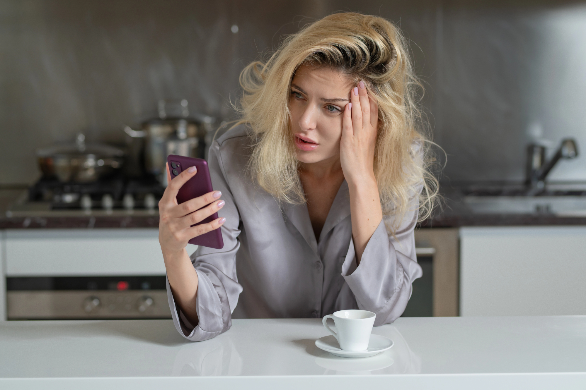 A woman with disheveled blonde hair looks worriedly at her smartphone while sitting at a kitchen counter. She wears a gray shirt and has a white coffee cup in front of her. In the background, there are kitchen appliances and pans.